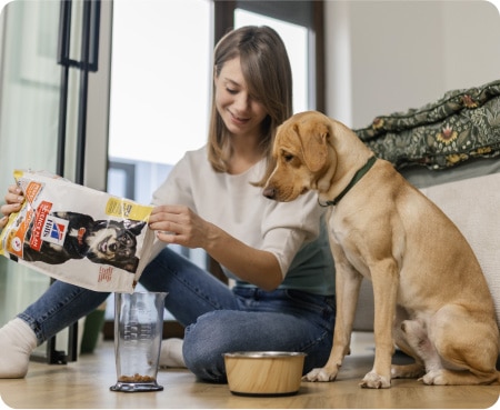 Жінка насипає корм «Hill's Pet Nutrition» у мірний контейнер, а собака уважно спостерігає за нею.